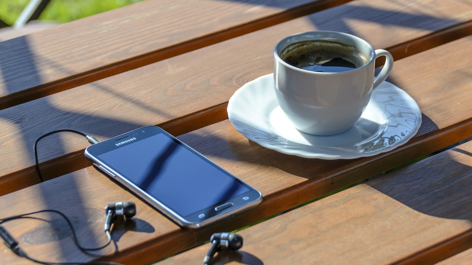 På ett soligt cafébord ligger en smartphone med inkopplade hörlurar bredvid fylld en kaffekopp.