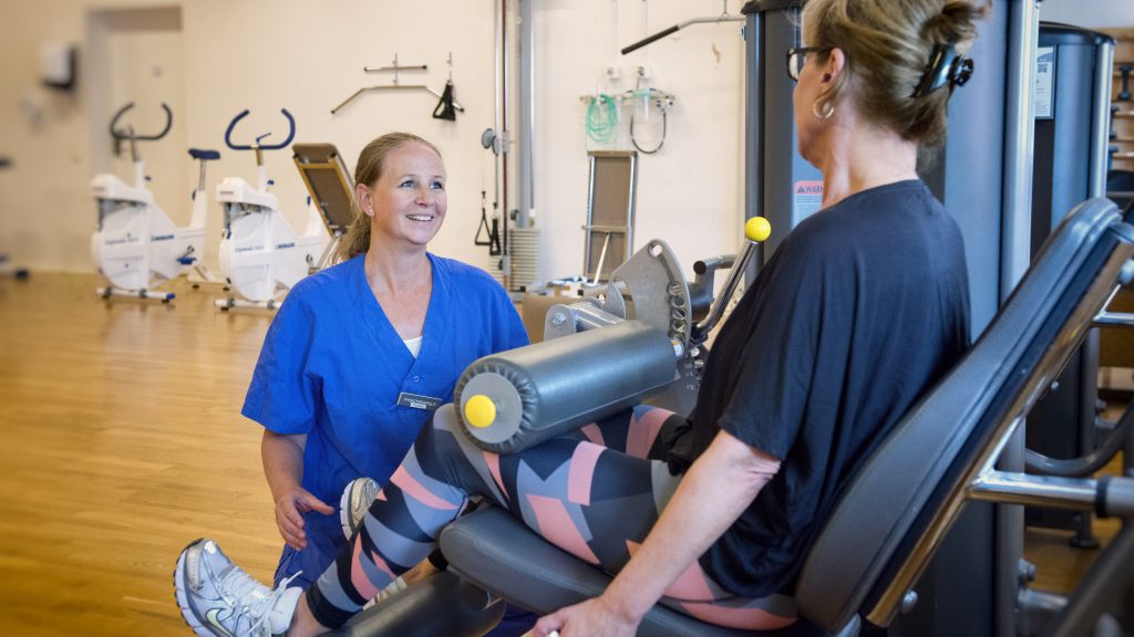 Fysioterapeuten Caroline pratar med en patient som sitter i en träningsmaskin