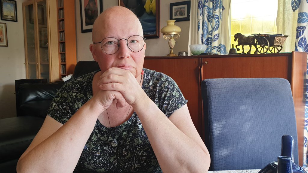 Bil på Lena som sitter vid ett köksbord med händerna knutna framför sig. Hon har inget hår på huvudet.