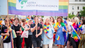 Bild från prideparaden där människor går med regnbågsflaggor och en skylt där det står Västra Götlandsregionen.