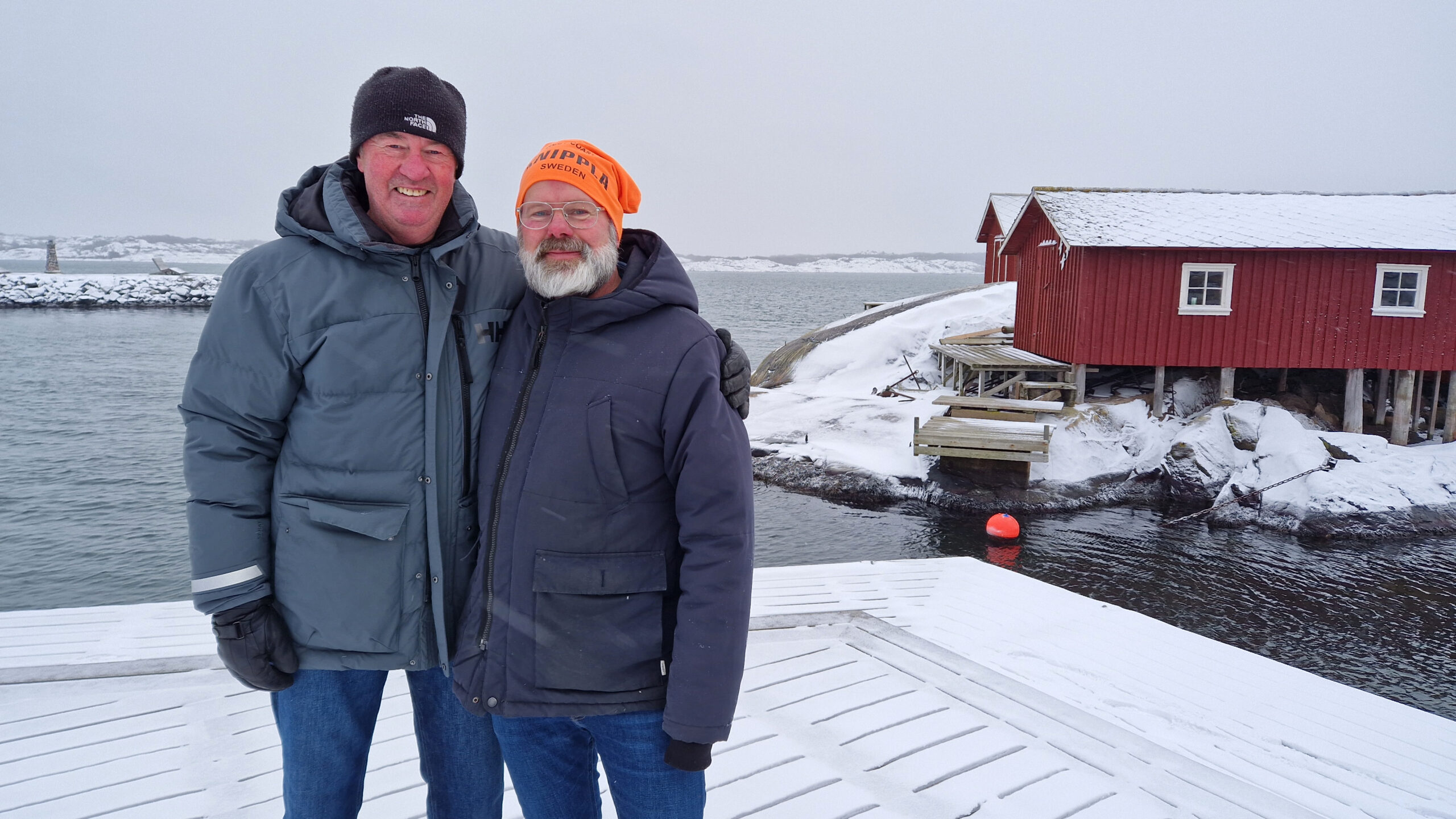 Två män står vid en kaj där man ser havet och sjöbodar i bakgrunden.