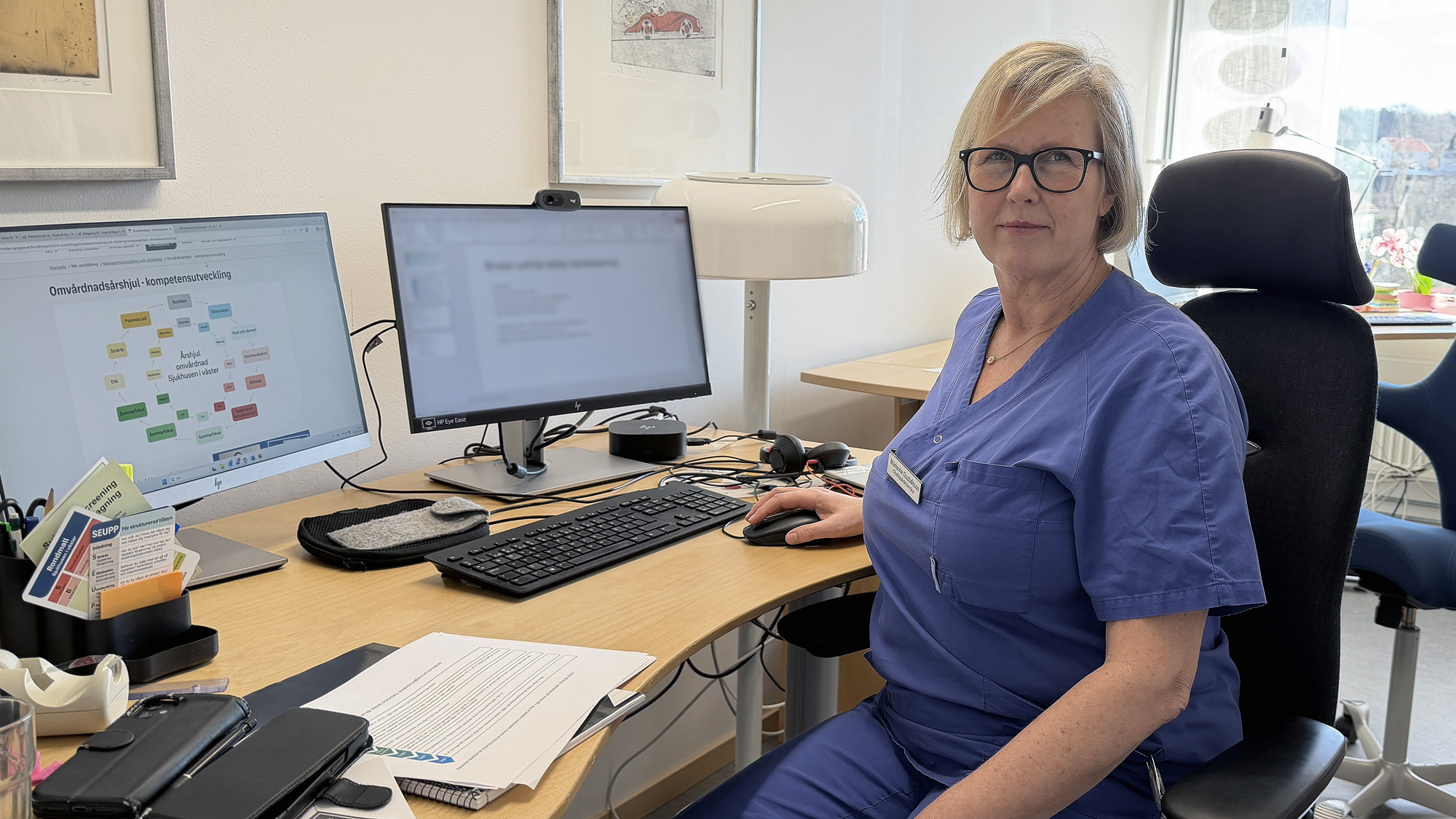 En kvinna i blå vårdklädsel sitter vid ett skrivbord med två datorskärmar i kontorsmiljö.