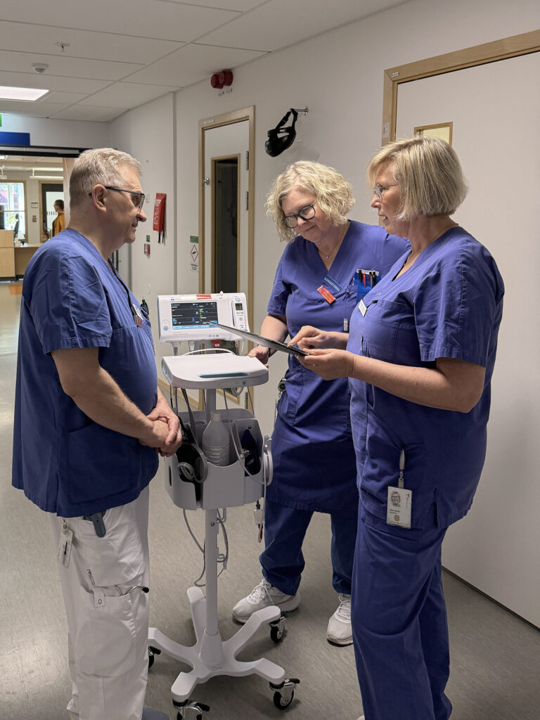 Tre personer i vårdklädsel står vid en apparat i en sjukhuskorridor och pratar med varandra.