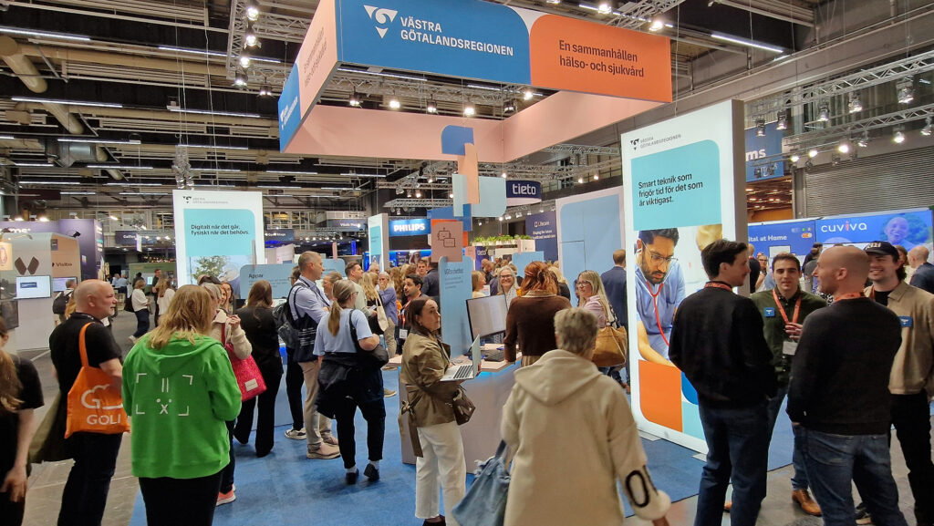 En stor grupp personer är samlade i och runt omkring en monter i en mässhall. Skyltar i taket med text om Västra Götalandsregionen.
